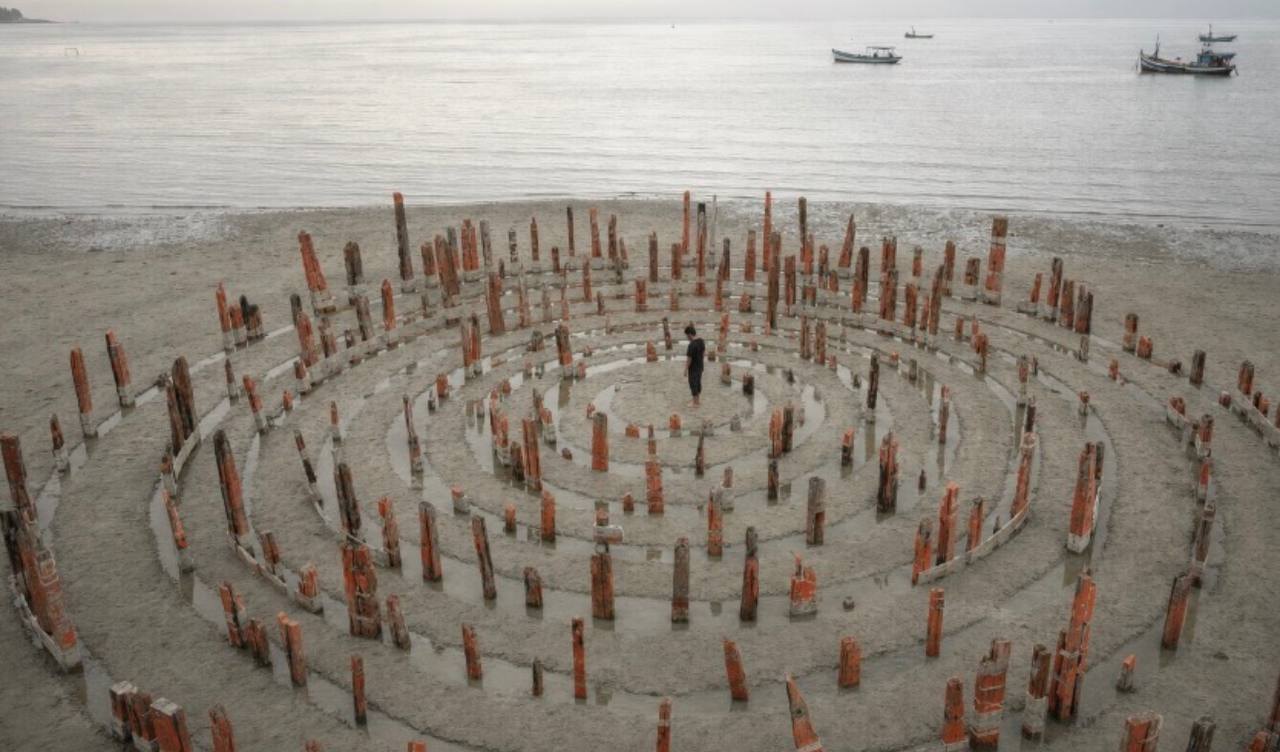 Concentric rings of standing posts on the beach with a figure at the centre